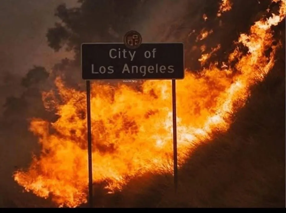 A black road sign reading 'City of Los Angeles' stands in front of a massive, roaring wildfire engulfing the background. The…