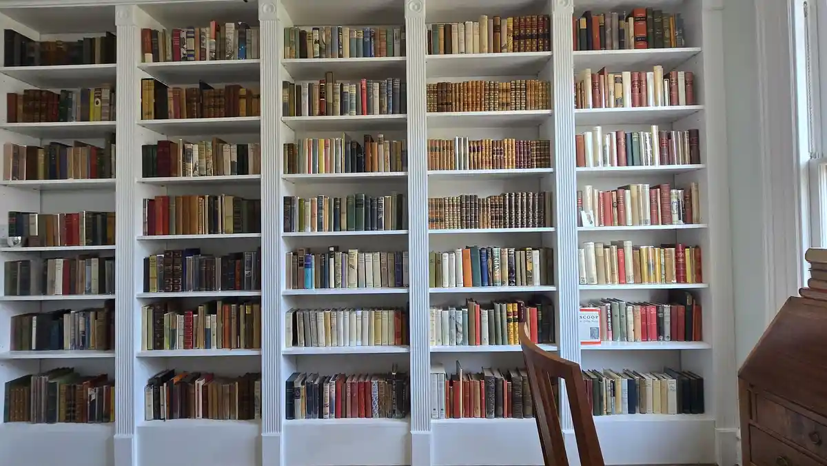 A photo of a large, white built-in bookshelf filled with numerous books of various sizes and colors, with a wooden chair…