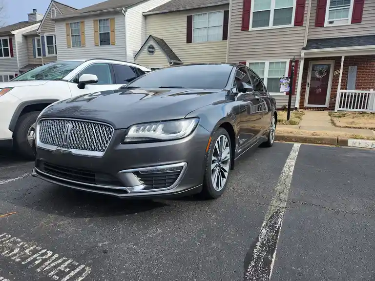 A dark gray Lincoln sedan is parked in a residential parking spot marked with faded white lines, next to a white SUV. Behind…