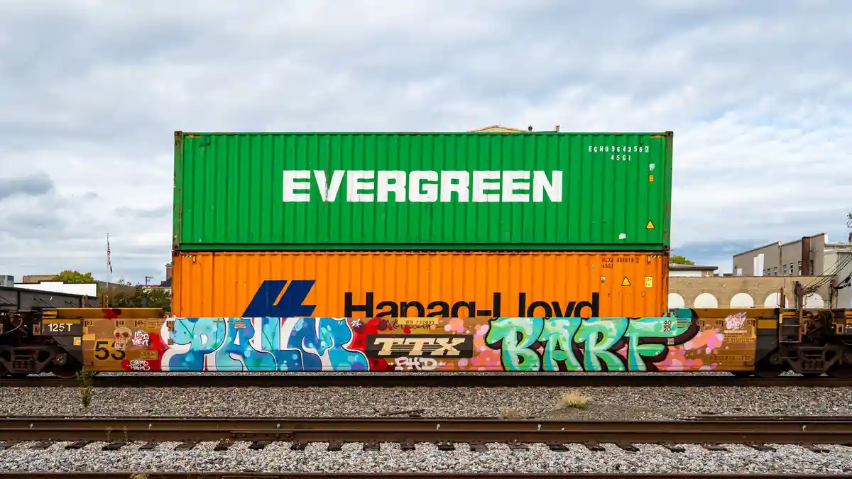 A green Evergreen shipping container is stacked atop an orange Hanjin-Lloyd container on a freight train car covered in…