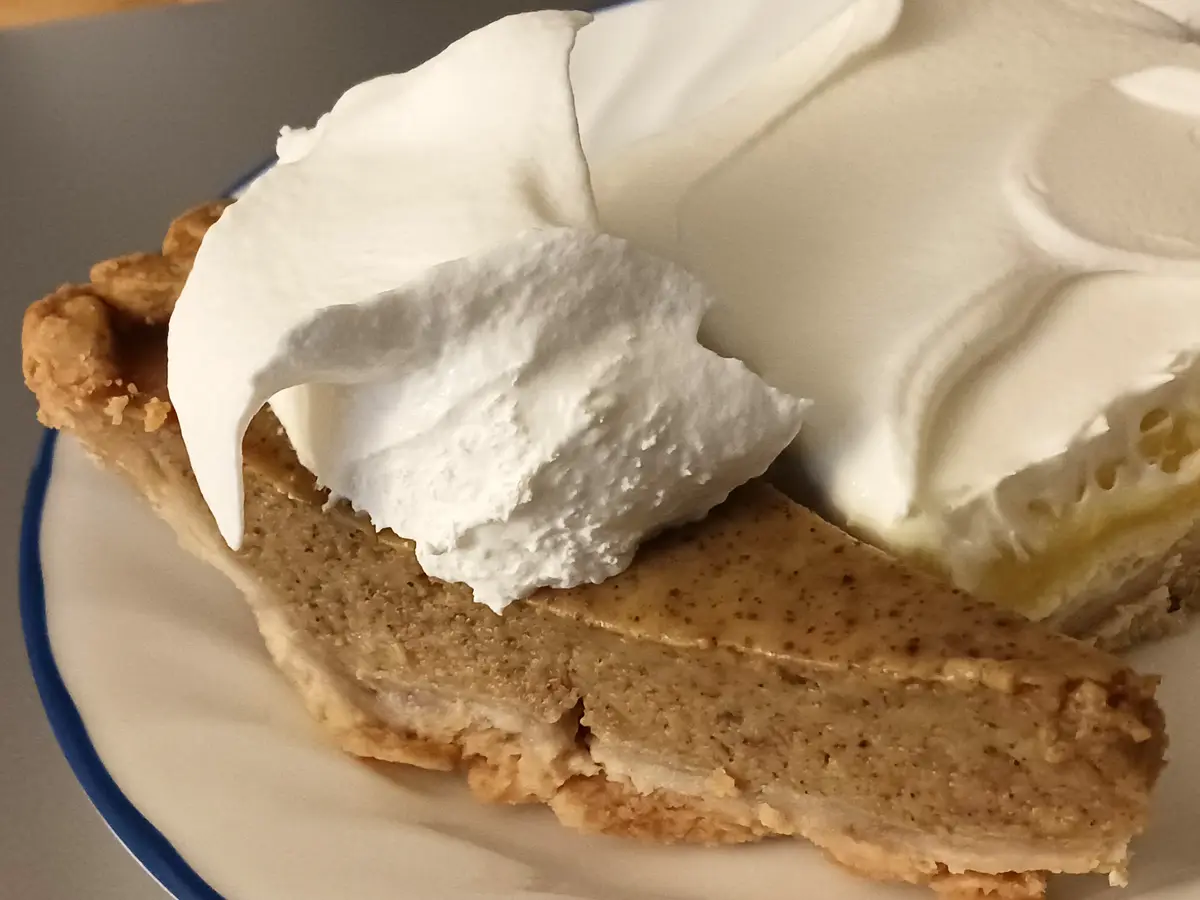 A close-up photo shows a slice of pie with a golden-brown crust and light brown filling, topped with a generous dollop of…