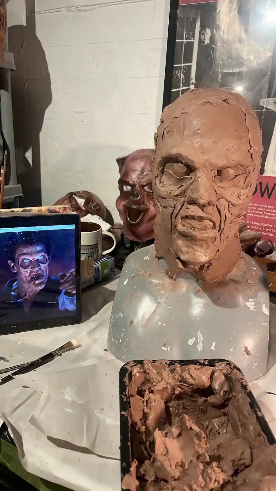 A work-in-progress zombie bust sculpture covered in brown clay sits on a messy table next to a container of clay, with a…