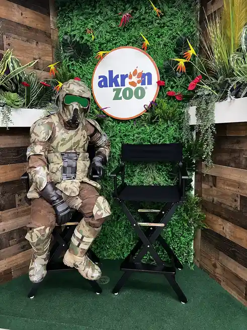 A person in full camouflage military gear, including a helmet with green-tinted goggles, sits on a black director's chair…