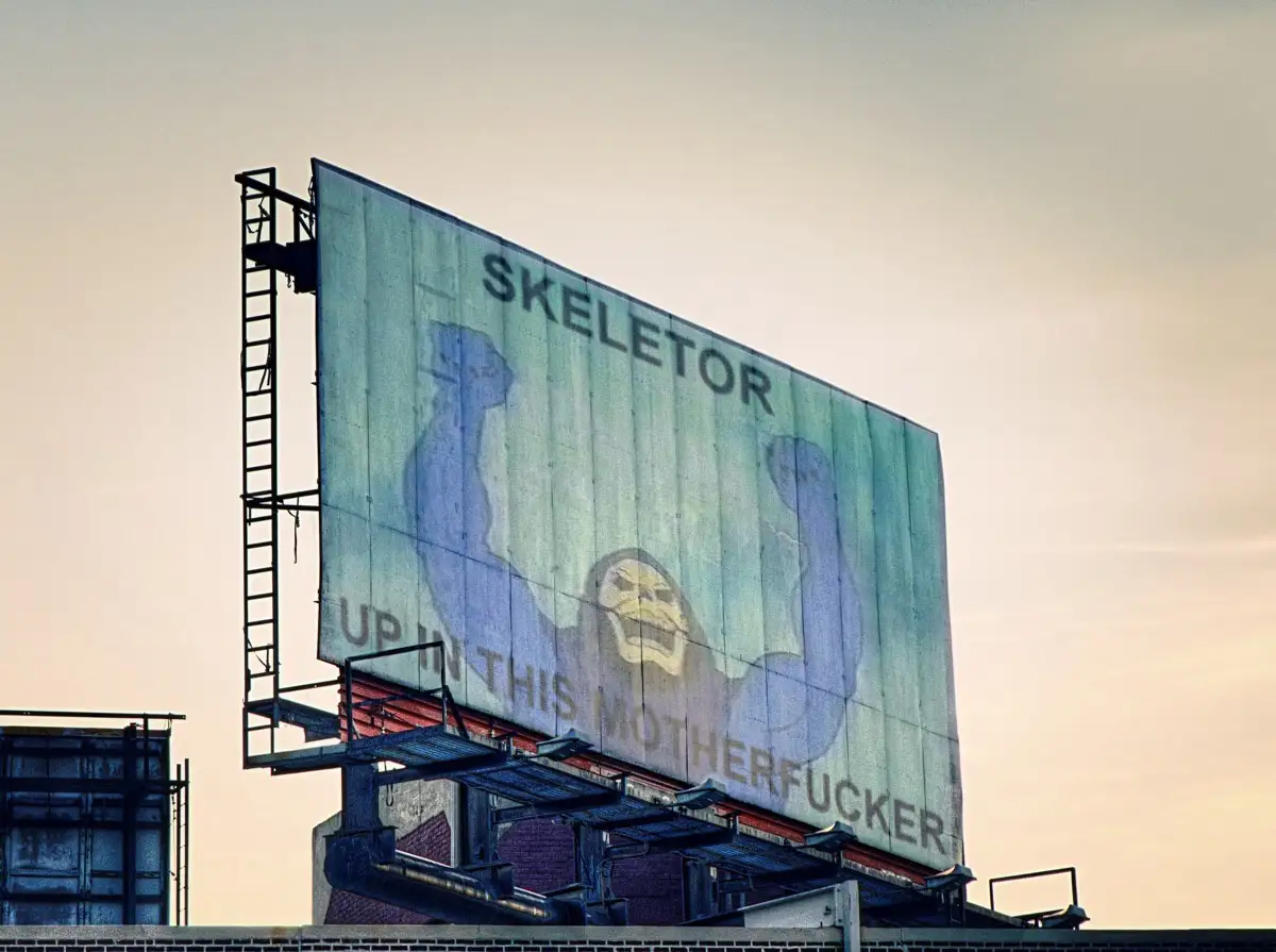 A weathered, large outdoor billboard displays a faded image of the character Skeletor from the Masters of the Universe…