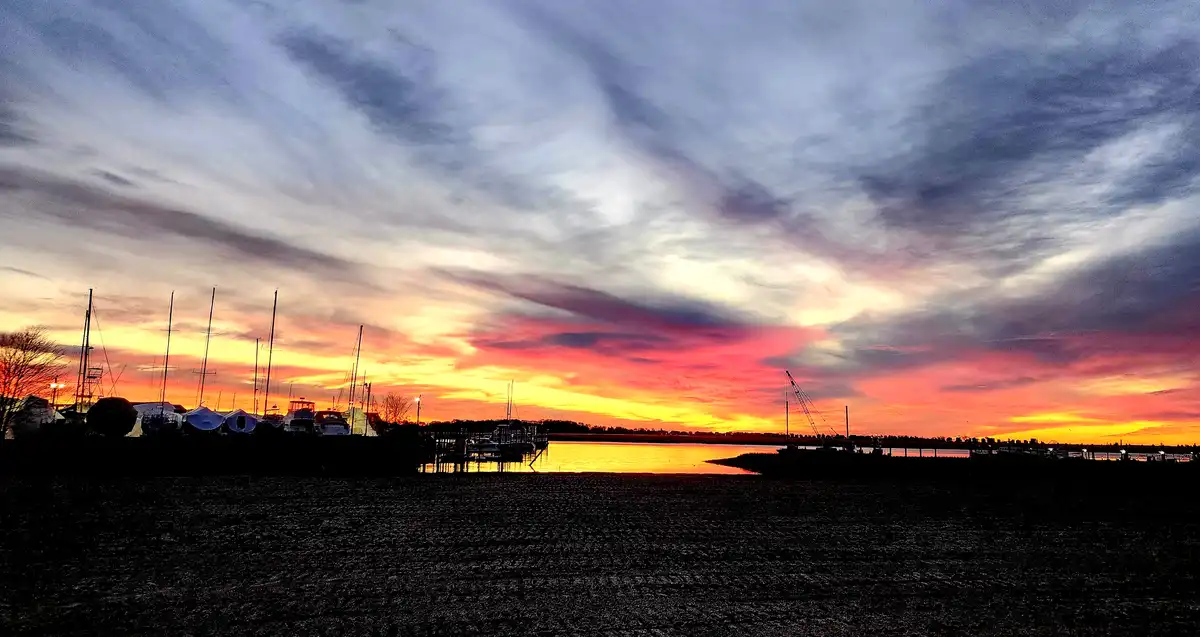 A scenic sunset over a marina with boats docked, silhouetted against vibrant orange, pink, and purple clouds. The foreground…