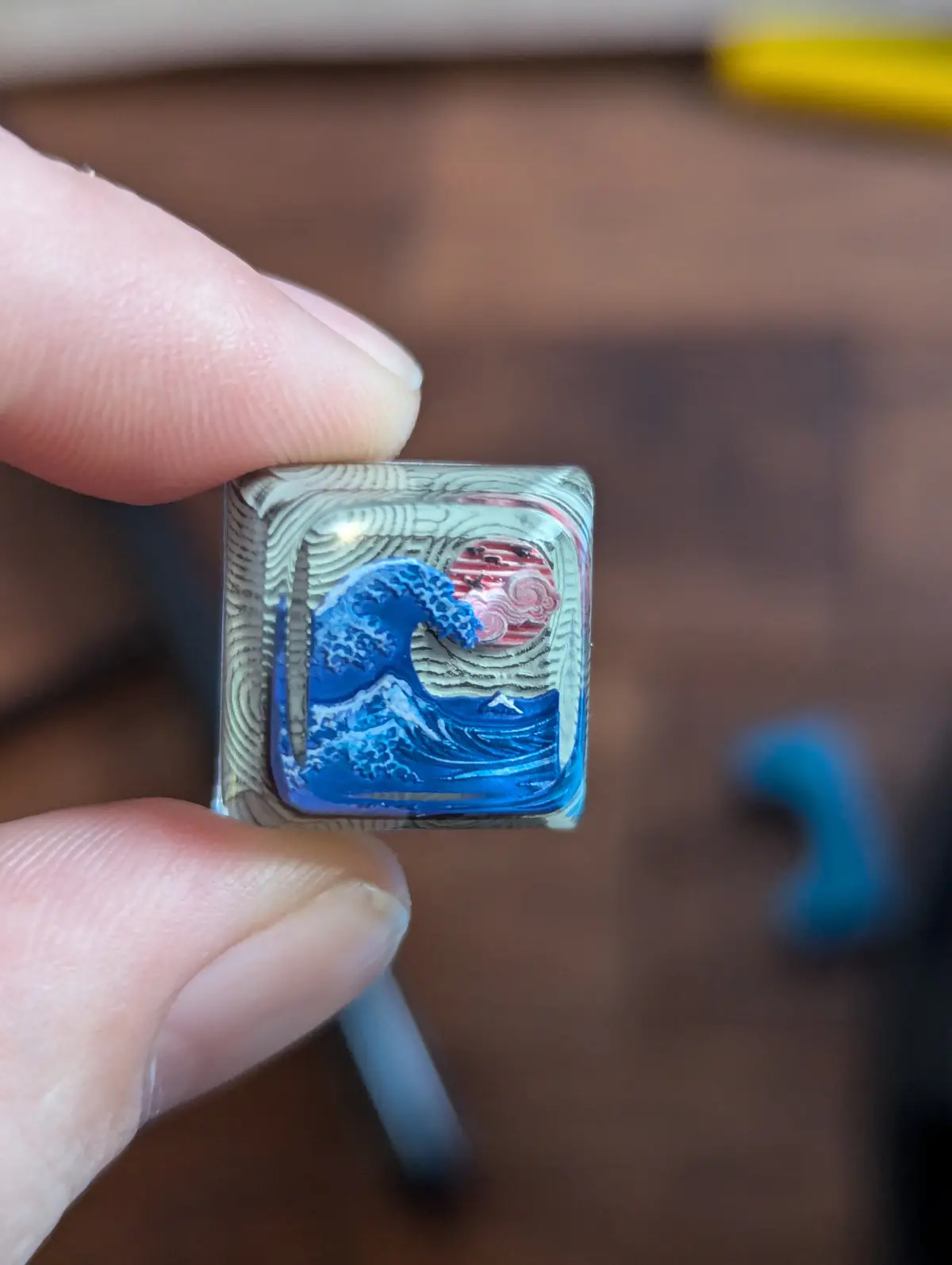 A hand holds a small, square, transparent object with a miniature, intricate painting of 'The Great Wave off Kanagawa'…