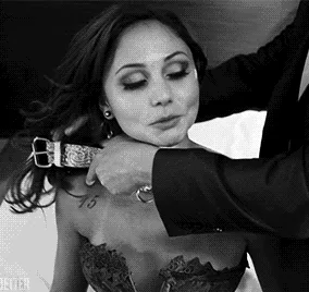 A black-and-white sequence shows a woman in a strapless dress as a man adjusts a choker around her neck. The number '15' is…