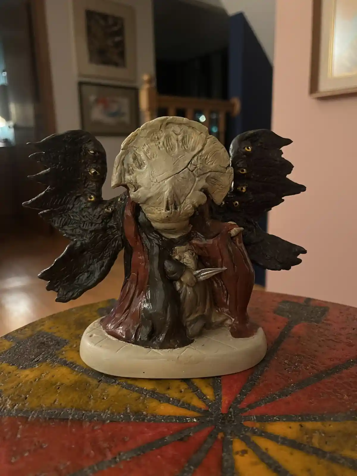 A figurine of a demonic-looking angel with a skull-like face and black wings, each wing hosting multiple small birds with…
