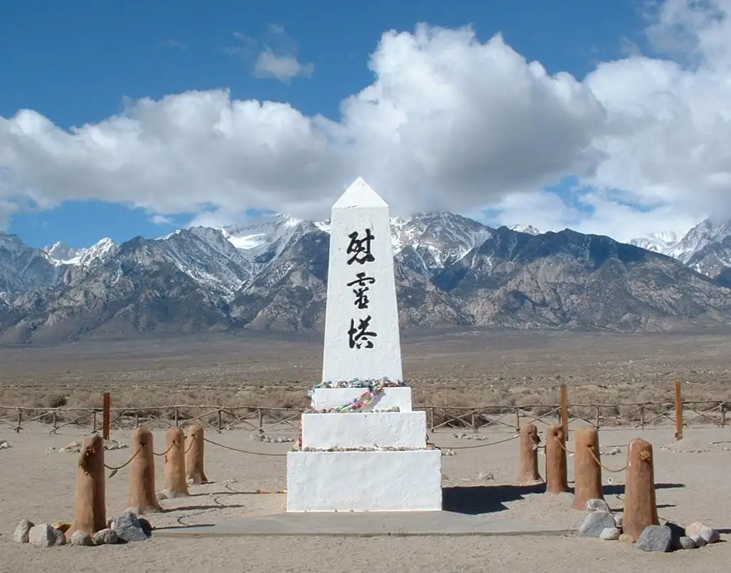 A white obelisk monument stands in a desert with snow-capped mountains in the background, surrounded by wooden posts and…