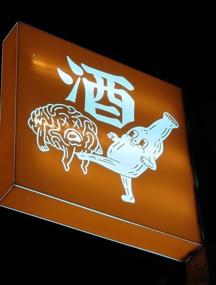 An illuminated orange sign at night displays a large white Chinese character '酒' (jiǔ, meaning alcohol) above a cartoonish…