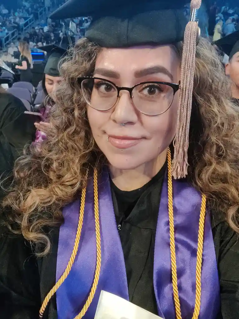 A young woman with curly hair and glasses wears a black graduation cap and gown with a purple and gold stole, smiling at the…