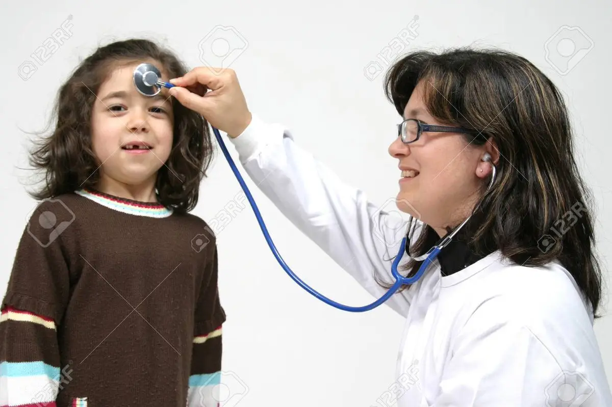 A woman in a white doctor's coat holds a blue stethoscope to a young girl's forehead while smiling. The girl, wearing a…