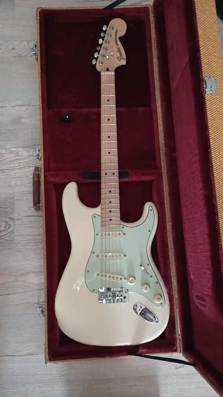 A Fender Stratocaster electric guitar with a cream body and light green pickguard rests inside an open, burgundy…