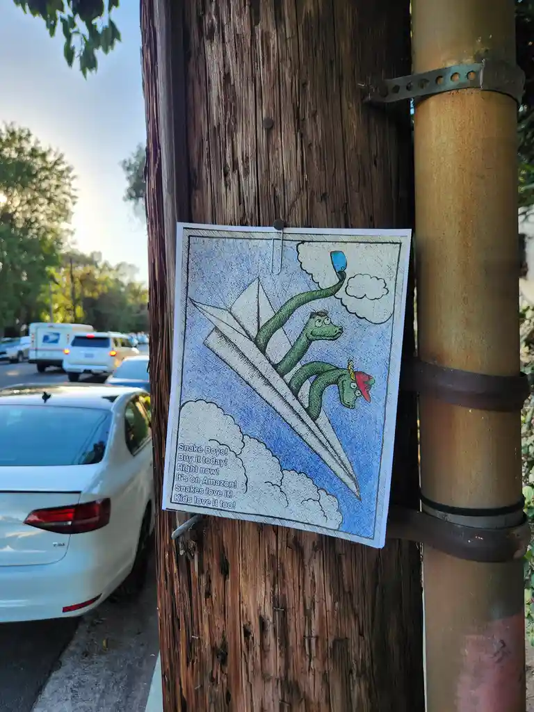 A hand-drawn, colored cartoon poster is clipped to a weathered wooden utility pole with a paperclip. The poster depicts two…