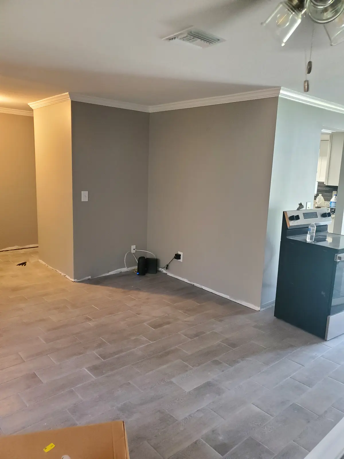 An empty room with gray walls, light wood-look flooring, and a ceiling fan, with a stove visible in the adjacent kitchen…