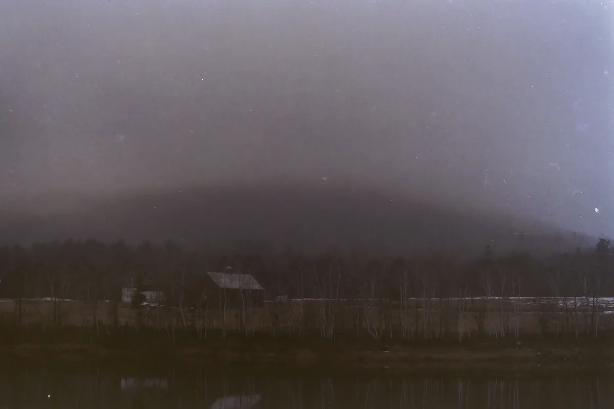 A grainy, vintage-style photograph shows a desolate, foggy landscape with bare trees, a small house, and a body of water in…