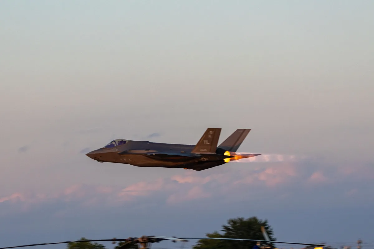A gray F-35 fighter jet is captured in mid-flight against a dusky sky with pink and blue clouds, its afterburner glowing…