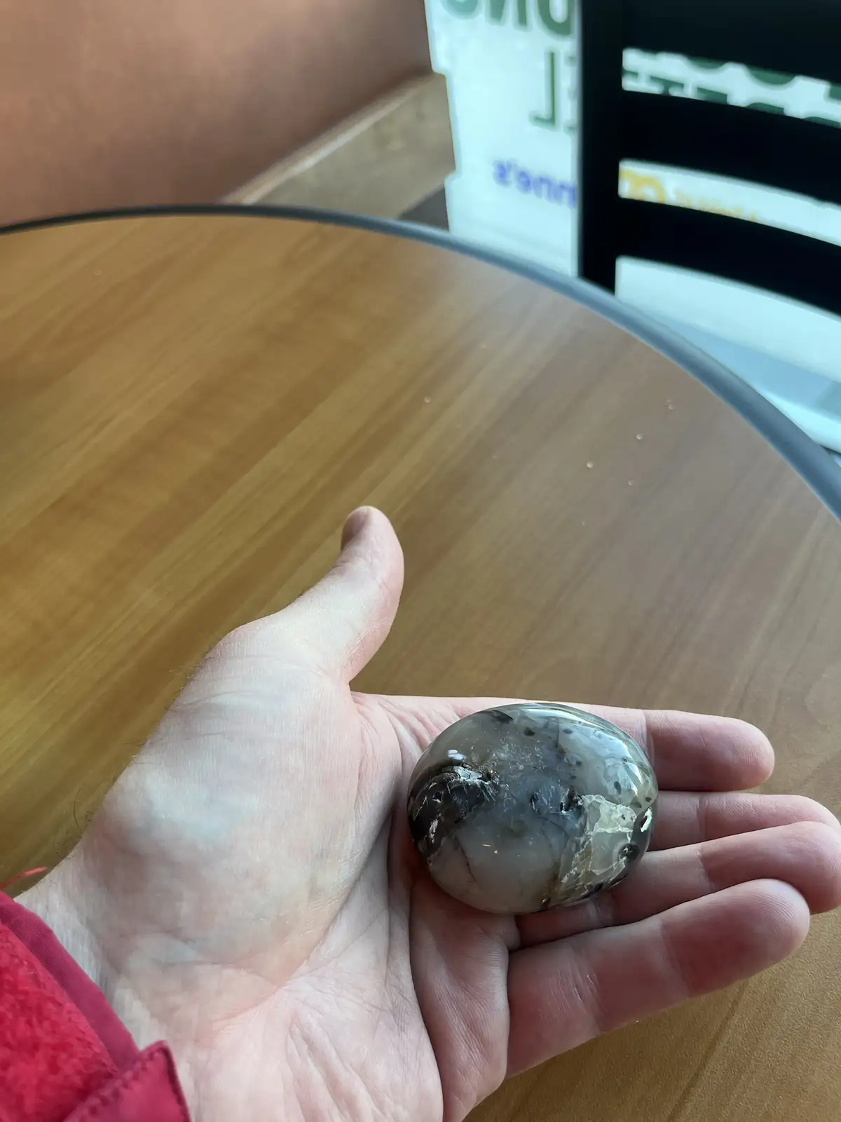 A person's hand holds a smooth, round, grayish stone with black and white inclusions over a wooden table; the overlaid text…