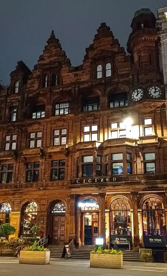 A nighttime photo of a grand, ornate, multi-story red sandstone building with arched windows and a prominent clock tower…