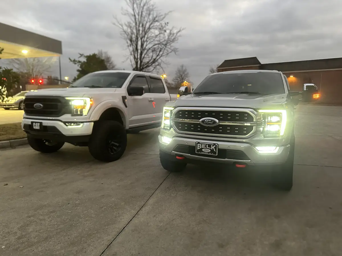 Two white Ford trucks are parked side by side in a parking lot at dusk, their headlights on. The truck on the left has a…