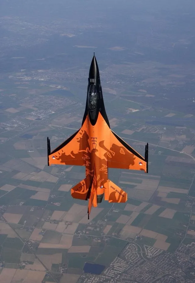 An orange and black fighter jet, viewed from above, flies over a patchwork landscape of fields and towns. The jet's tail fin…