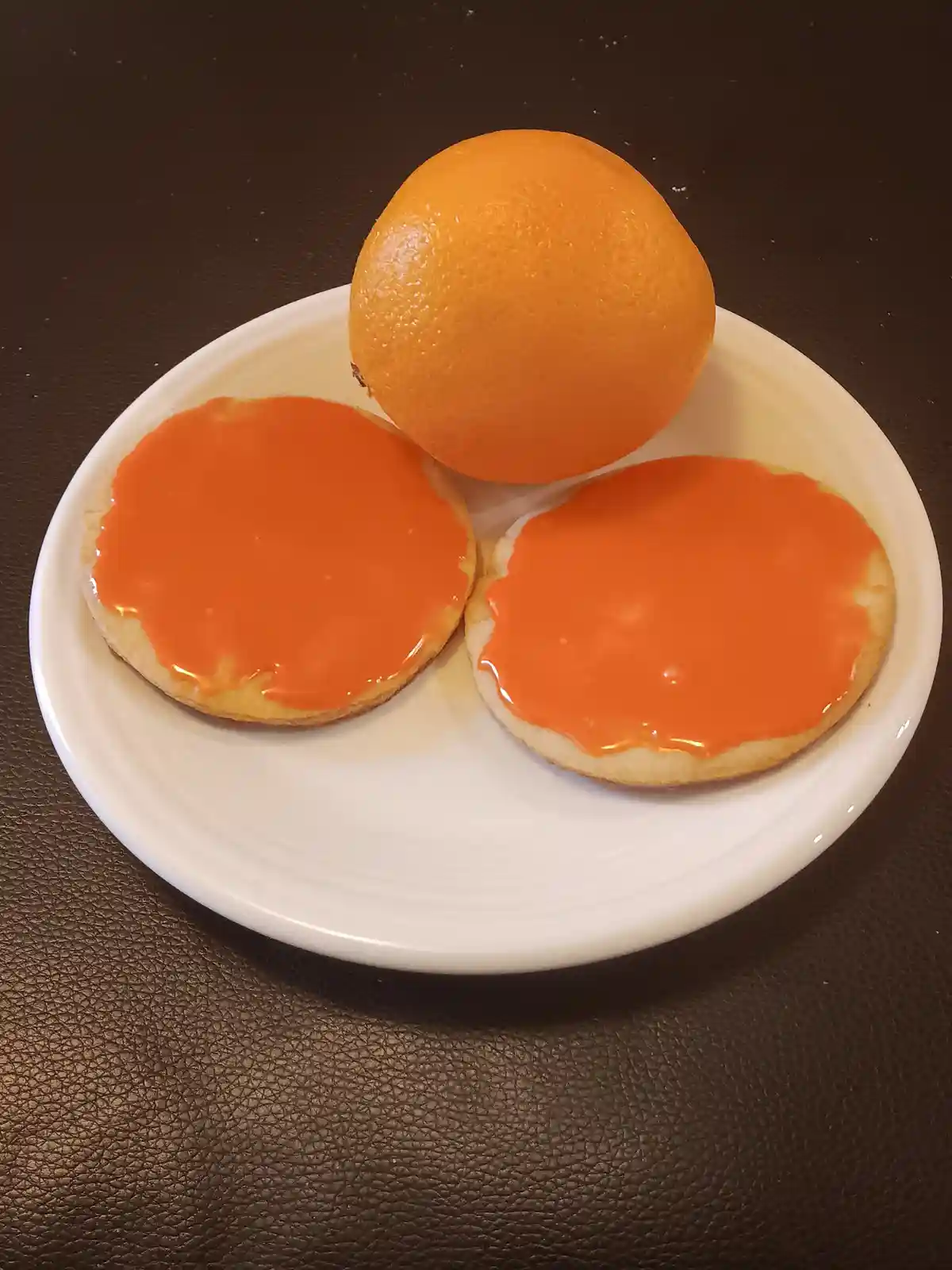 A white plate holds two round cookies topped with bright orange icing and a whole orange, arranged to visually mimic a face…
