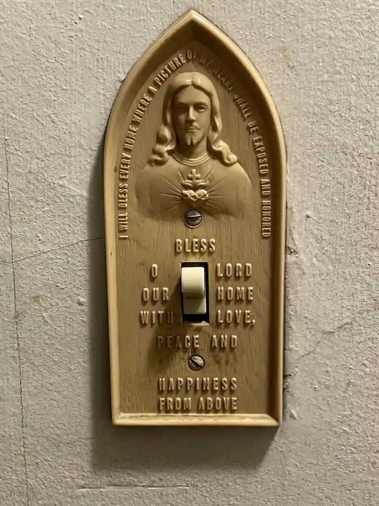 A beige, arched light switch plate features a raised relief of Jesus Christ with a Sacred Heart, surrounded by religious…