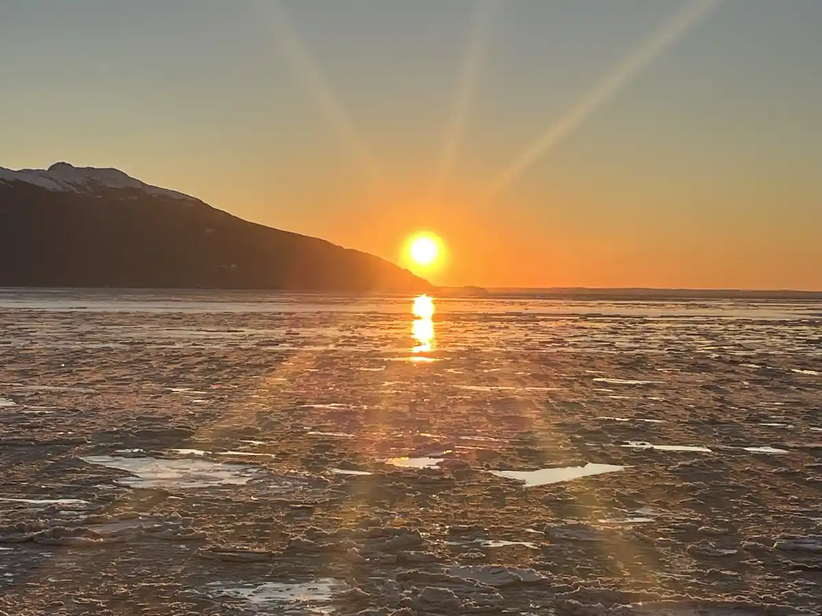 A serene sunset over a frozen body of water with broken ice floes reflecting the golden sun, a snow-capped mountain…
