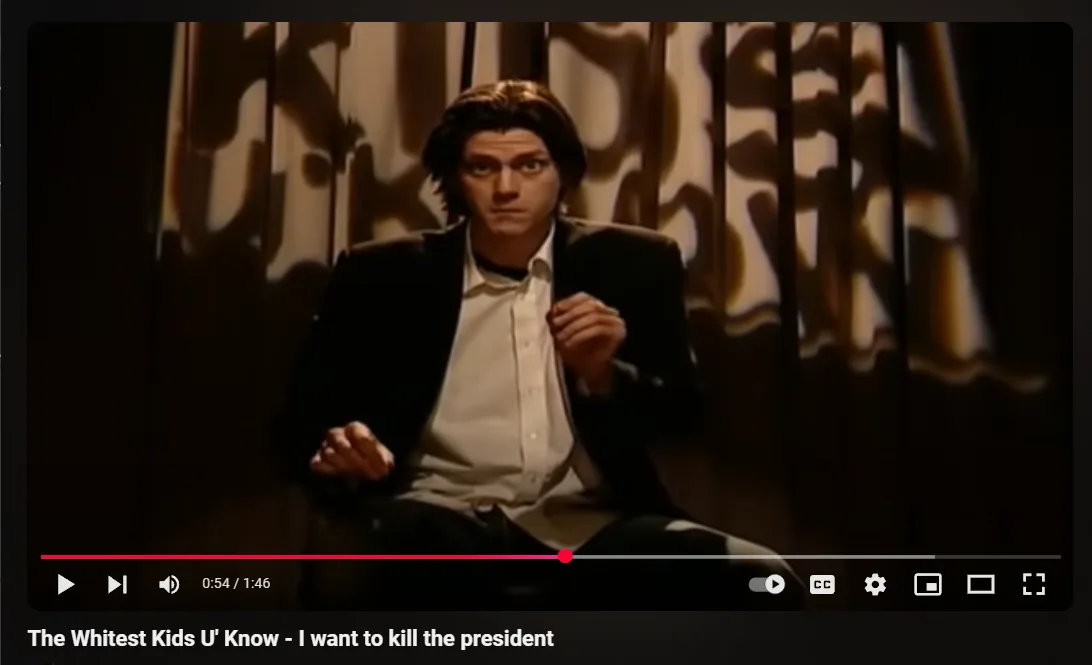 A still from a music video shows a young man in a dark suit and white shirt sitting with a serious, intense expression, his…