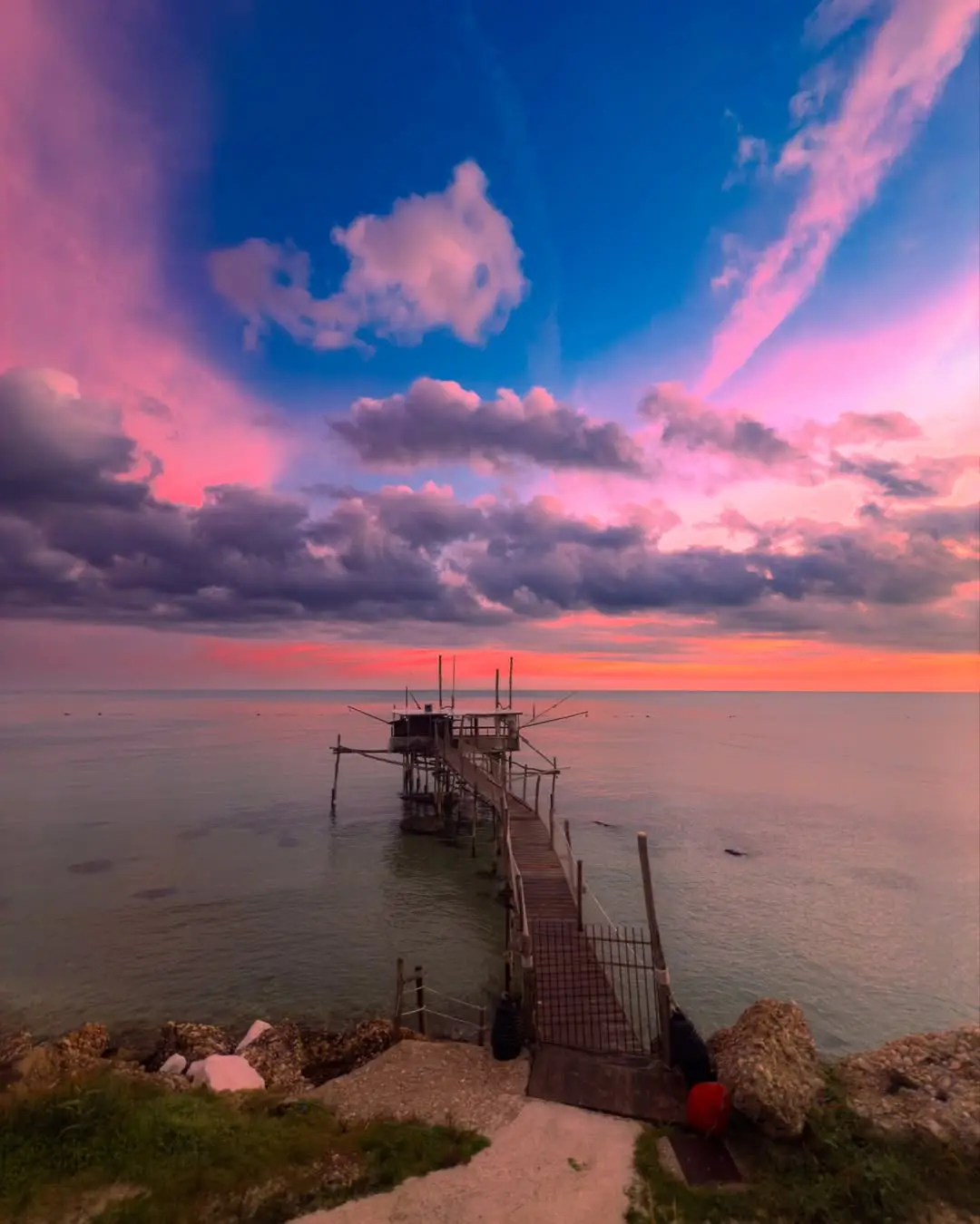 A serene sunset photograph shows a wooden pier extending into calm water under a vibrant sky of pink, purple, and orange…