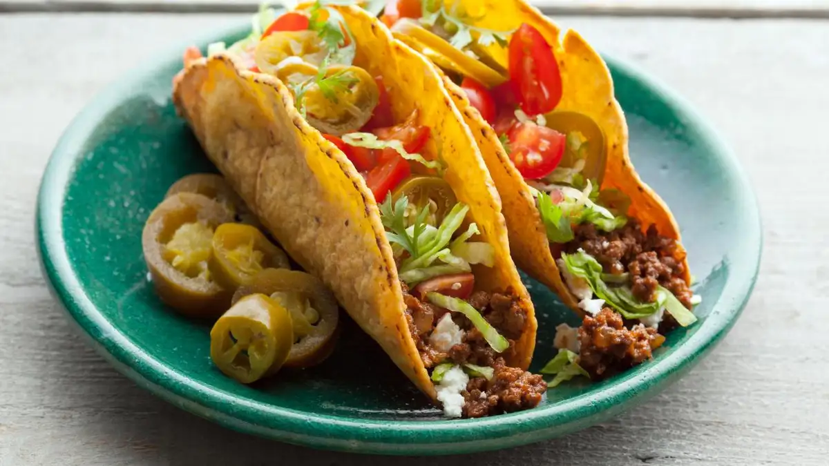 Three soft tacos filled with ground meat, lettuce, tomatoes, and cilantro sit on a green plate alongside sliced jalapeños…