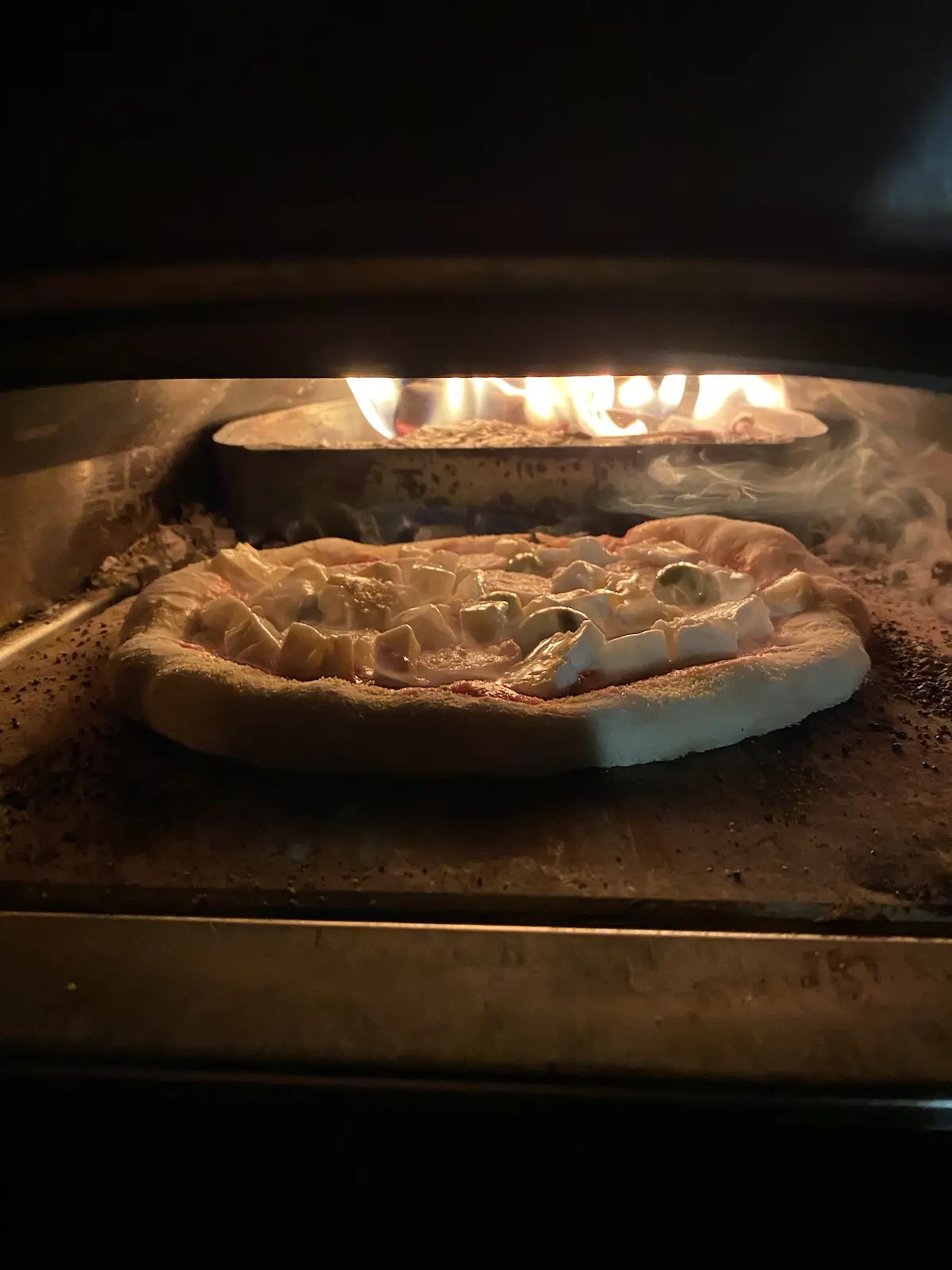 A close-up shot from inside a wood-fired oven shows a pizza baking on a stone surface, with visible flames and smoke rising…