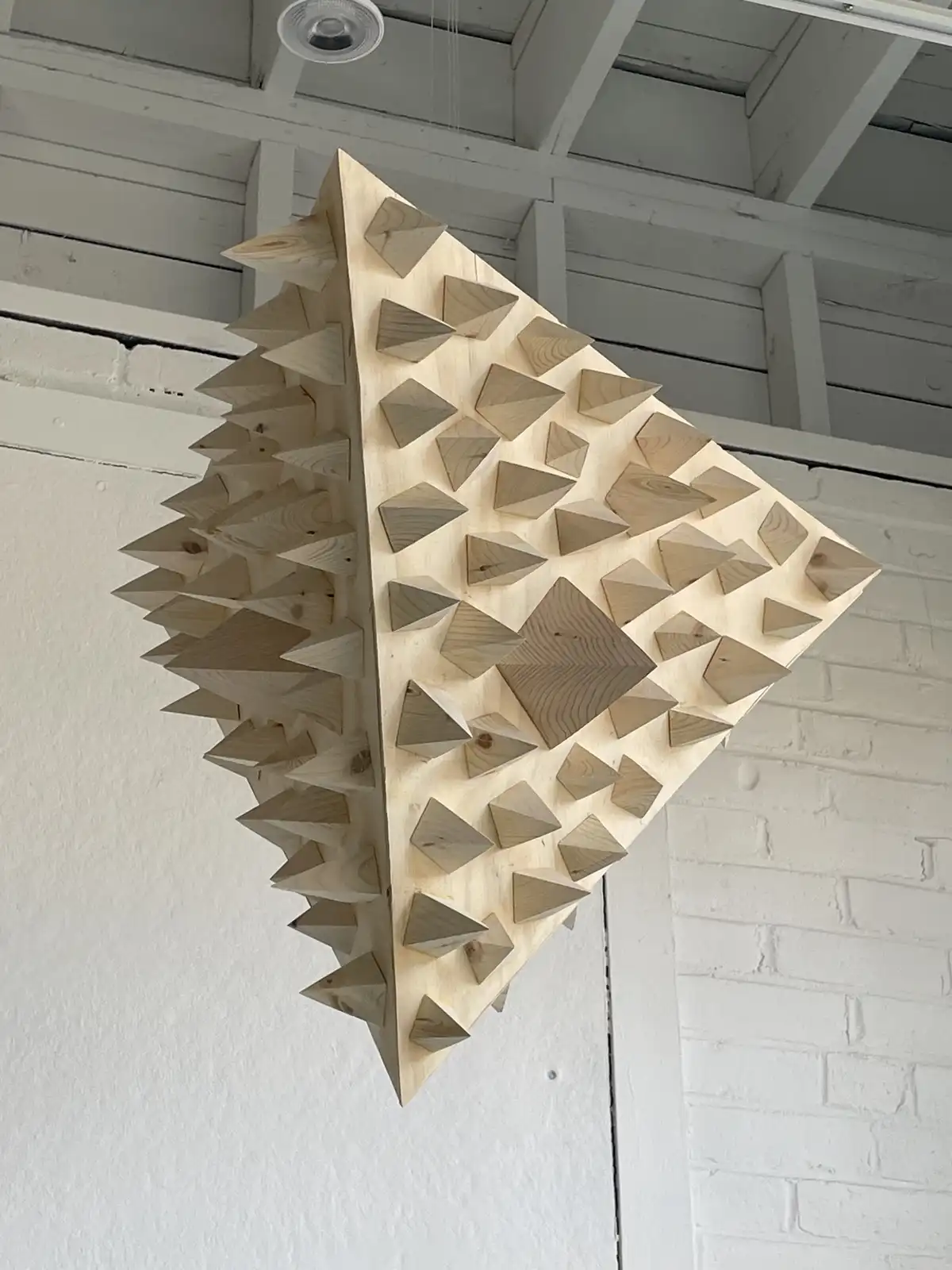 A wooden sculpture shaped like a diamond or pyramid, covered in numerous small, protruding wooden spikes, hangs from the…