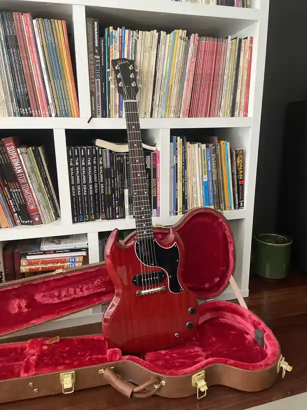 A red Gibson electric guitar sits upright inside its open, red-lined hard case, positioned in front of a white bookshelf…