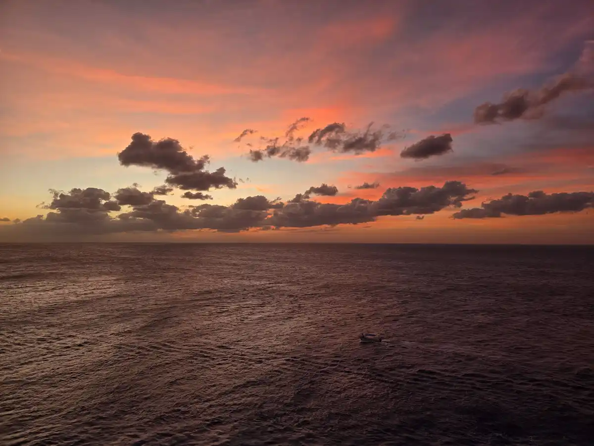 A serene aerial view of a vast ocean at sunset, with vibrant orange and purple clouds above and a small boat moving across…