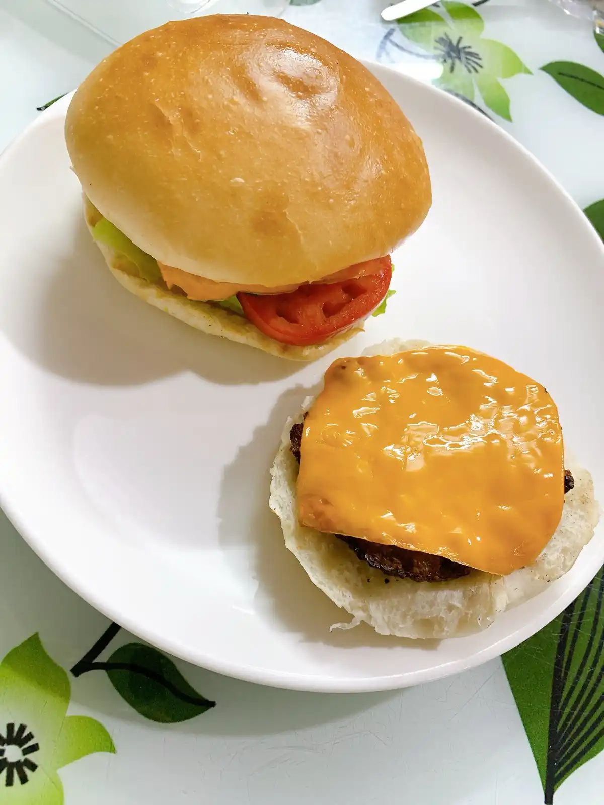 A white plate holds two halves of a cheeseburger; one half has the top bun, tomato, and lettuce, while the other half has…