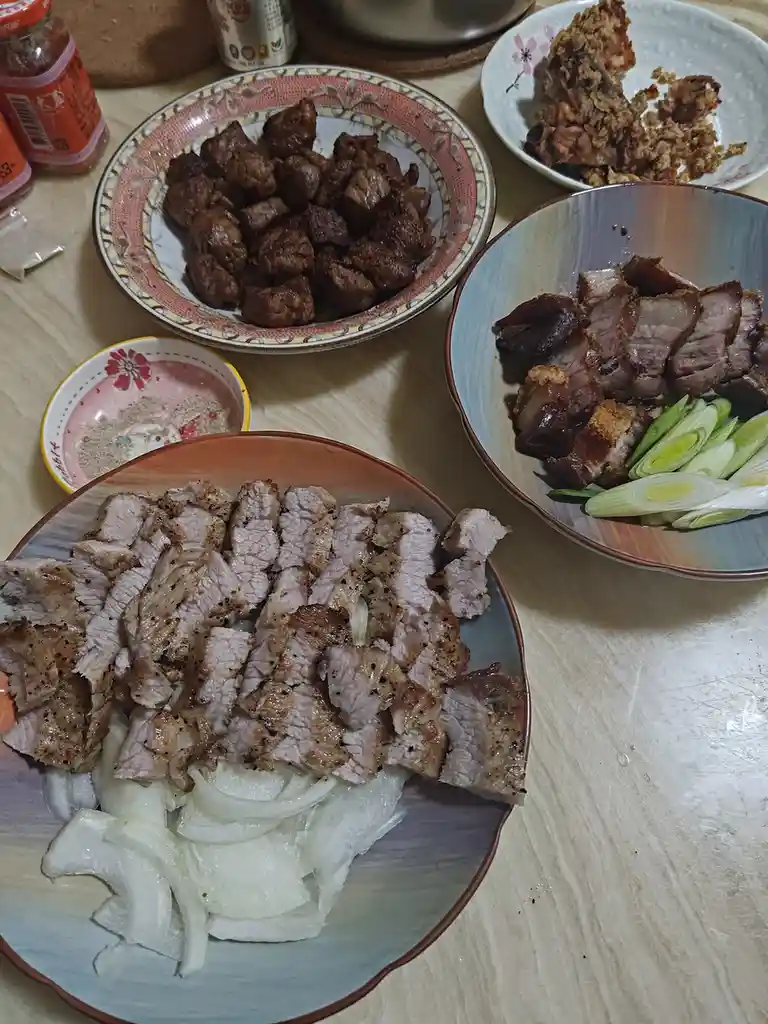 A table displays several plates of cooked meat dishes, including sliced grilled pork with onions, roasted meat with green…