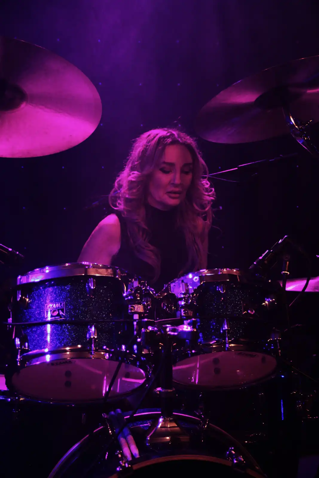 A woman with long blonde hair sits behind a glittery black Tama drum kit under purple stage lights, looking down with a…