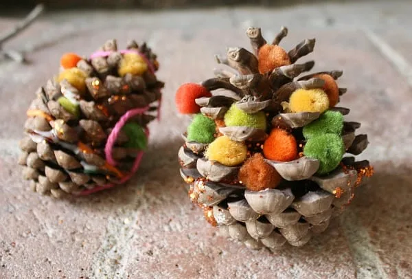Two decorated pinecones sit on a textured surface, adorned with colorful pom-poms, glitter, and yarn. The larger pinecone on…
