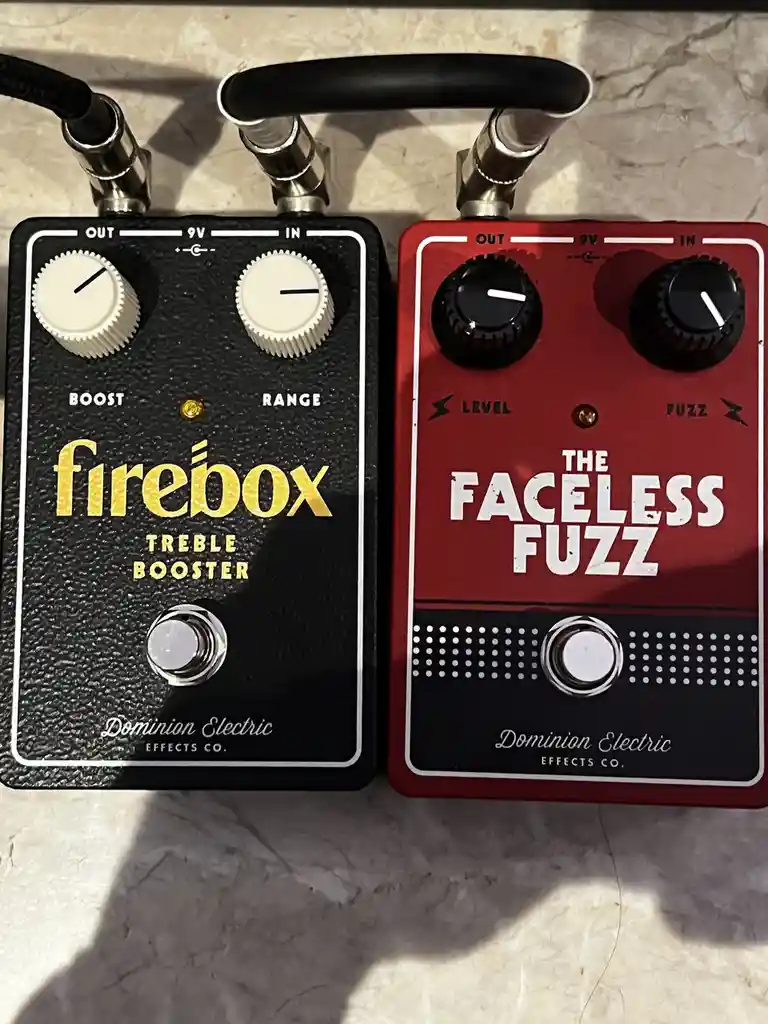 Two Dominion Electric guitar effects pedals sit side-by-side on a marble surface: a black 'firebox treble booster' and a red…