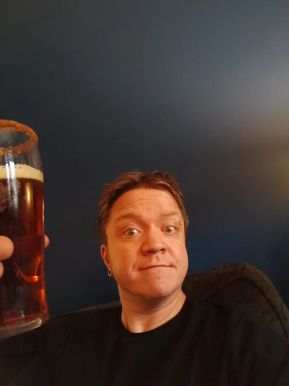 A person with light brown hair and multiple facial piercings holds up a tall glass of amber-colored beer with a foamy head…