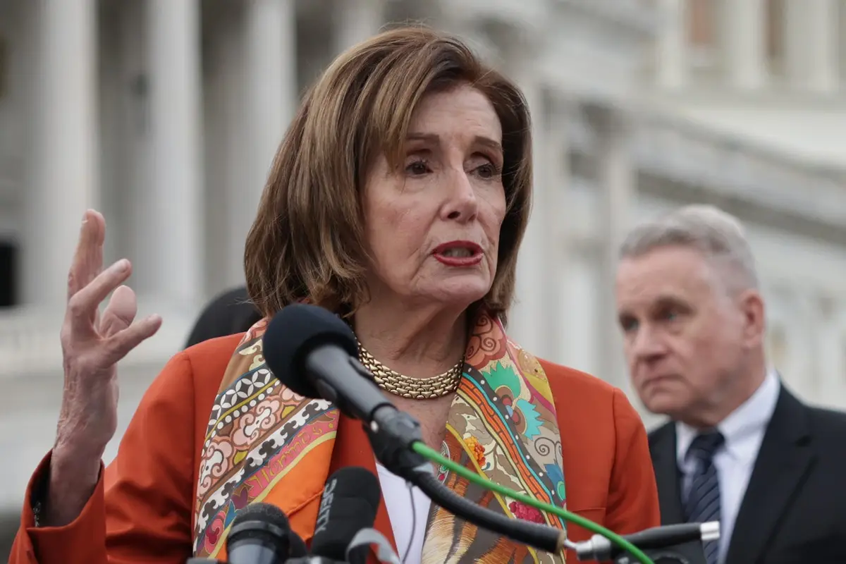A woman with brown hair wearing an orange jacket and patterned scarf speaks into microphones while gesturing with her right…