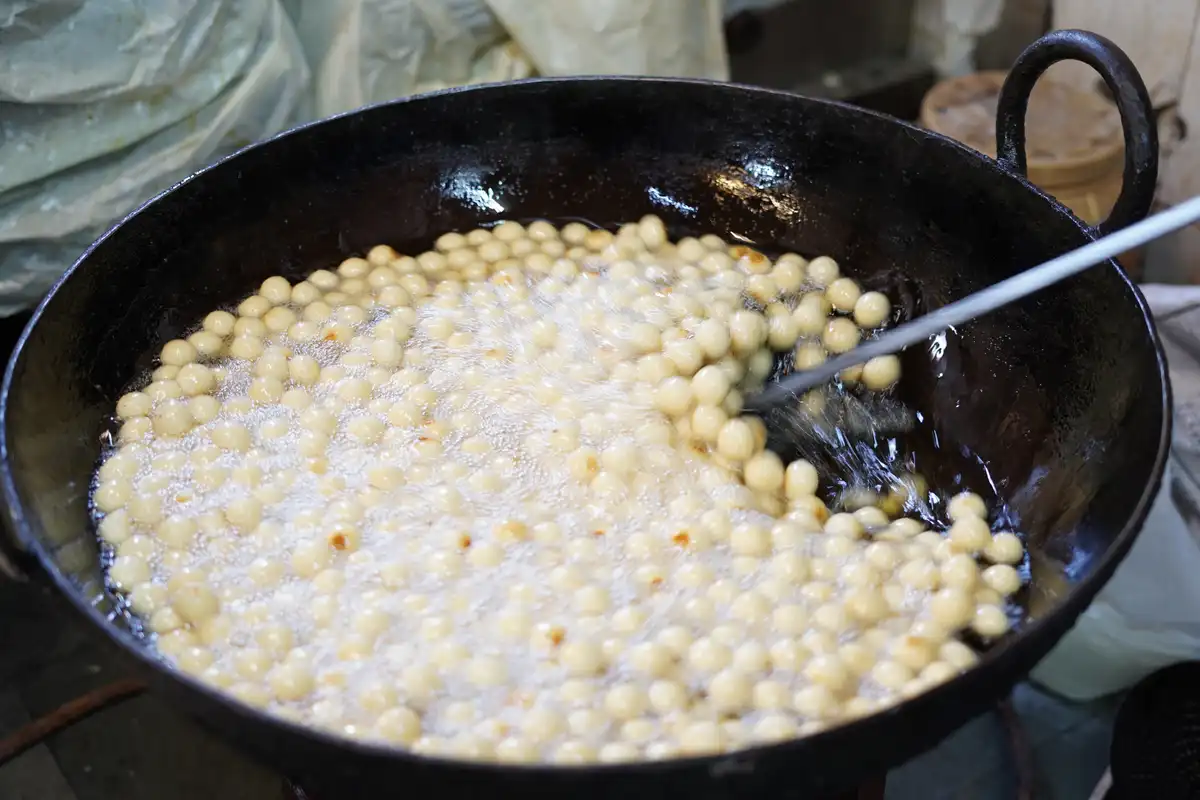 A large black wok filled with hot oil and numerous small, round, pale-yellow dough balls being fried, with a long white…