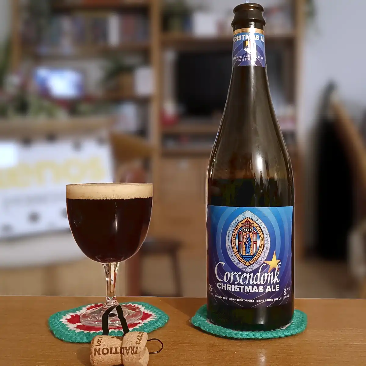 A bottle of Corsendonk Christmas Ale and a glass of the dark beer sit on green coasters on a wooden table, with a cork…