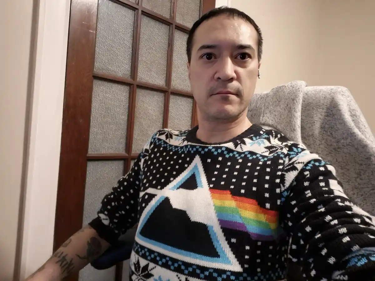 A man with short dark hair and a tattooed left arm sits in a chair, wearing a black, white, and blue knitted sweater…