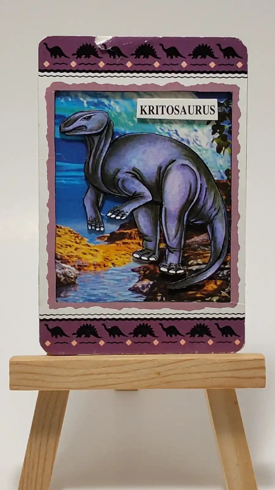 A vintage-style dinosaur card featuring a purple Kritosaurus standing near water, framed with purple borders and black…