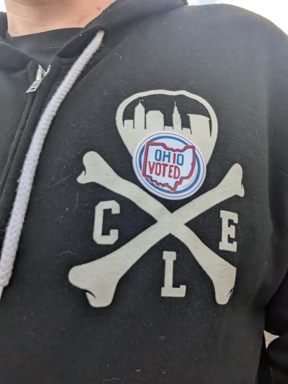 A close-up of a black zip-up hoodie with a large white skull and crossbones graphic. The skull's outline forms a city…
