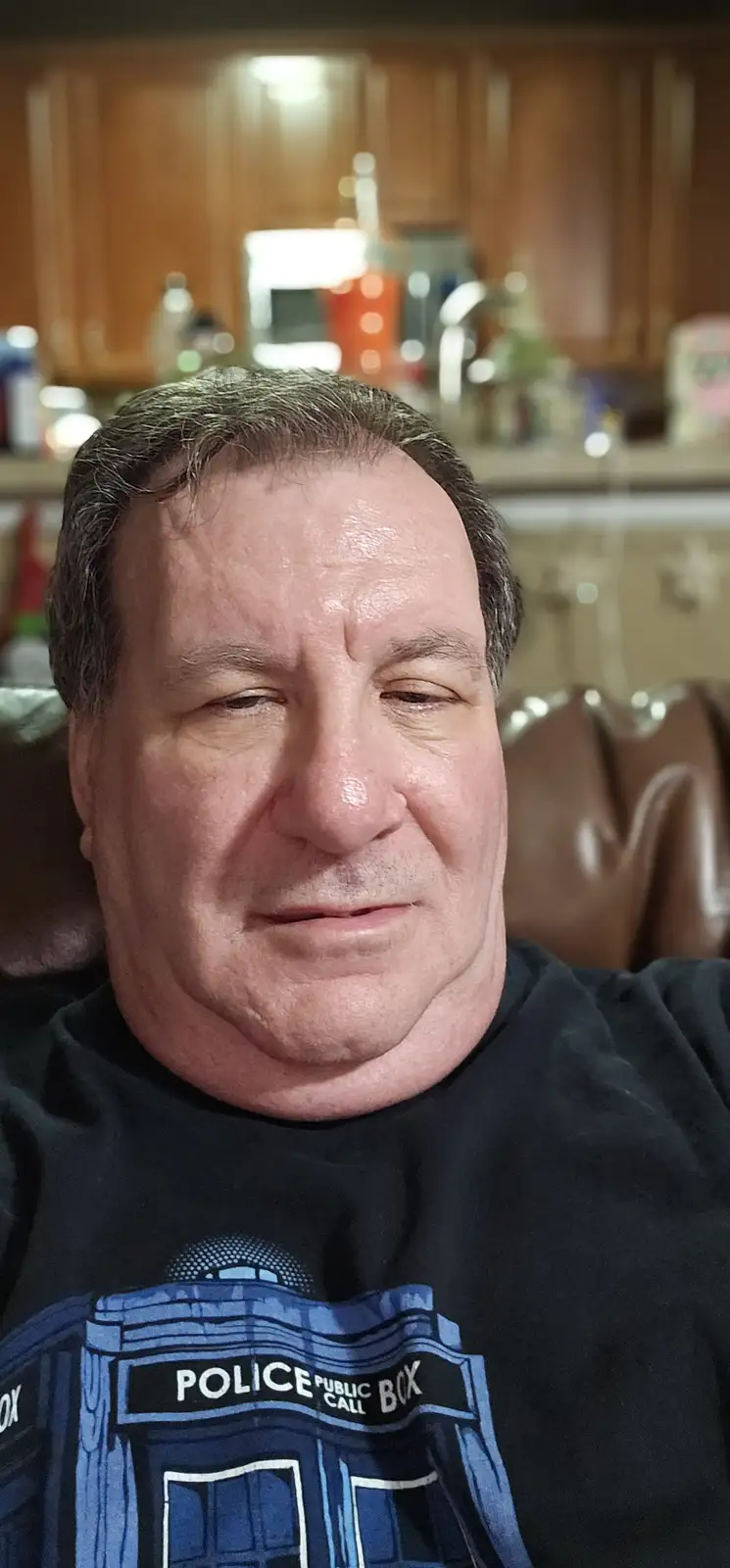 A man with a sweaty, tired expression sits in a brown leather chair in a kitchen, wearing a black t-shirt with a blue police…