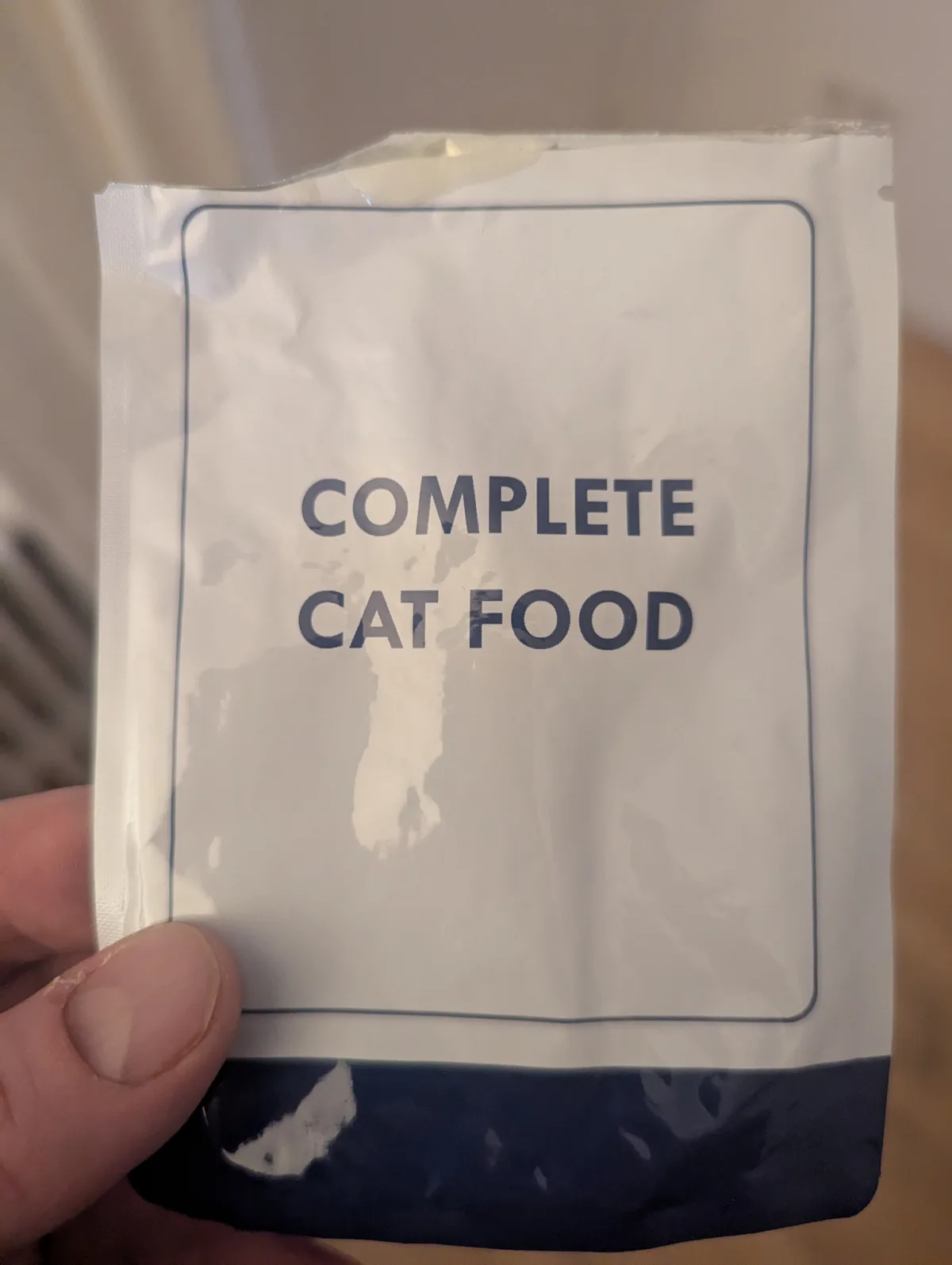 A hand holds a white, crinkled plastic packet with bold, dark blue text reading 'COMPLETE CAT FOOD' inside a thin blue…