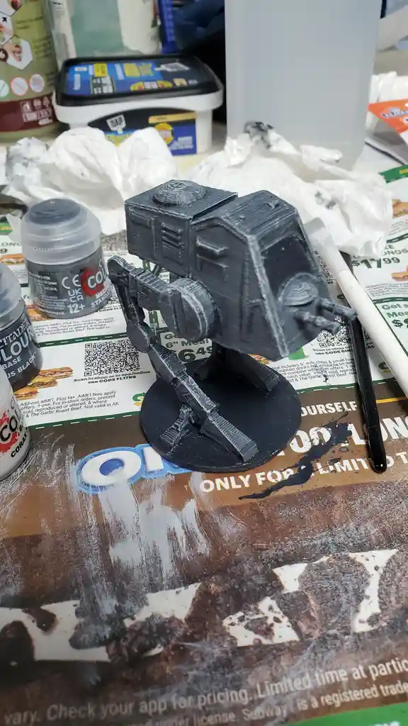 A small, unpainted AT-AT walker model sits on a black base atop a Subway restaurant table, surrounded by paint bottles and…